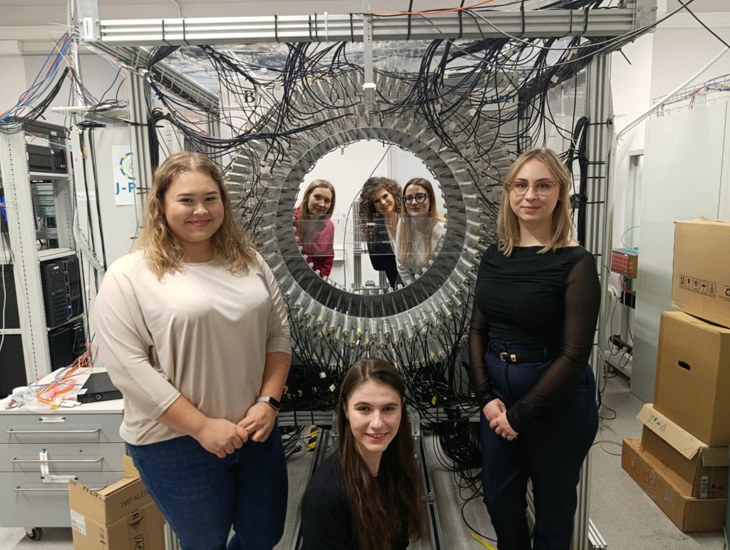 Studentki wraz z opiekunką wizyty Panią dr inż. Mileną Piotrowską pozują do zdjęcia przy specjalistycznym urządzaniu w laboratorium J-PET. 