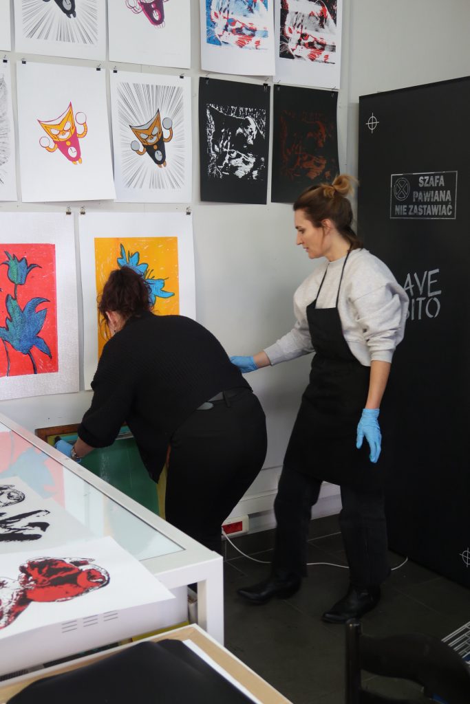 Dwie kobiety w czarnych fartuchach ochronnych pracują przy ścianie z ekspozycją prac. Jedna z nich pochyla się, operując przy ramie sitodrukowej, druga obserwuje proces. Obie mają założone niebieskie rękawiczki ochronne.