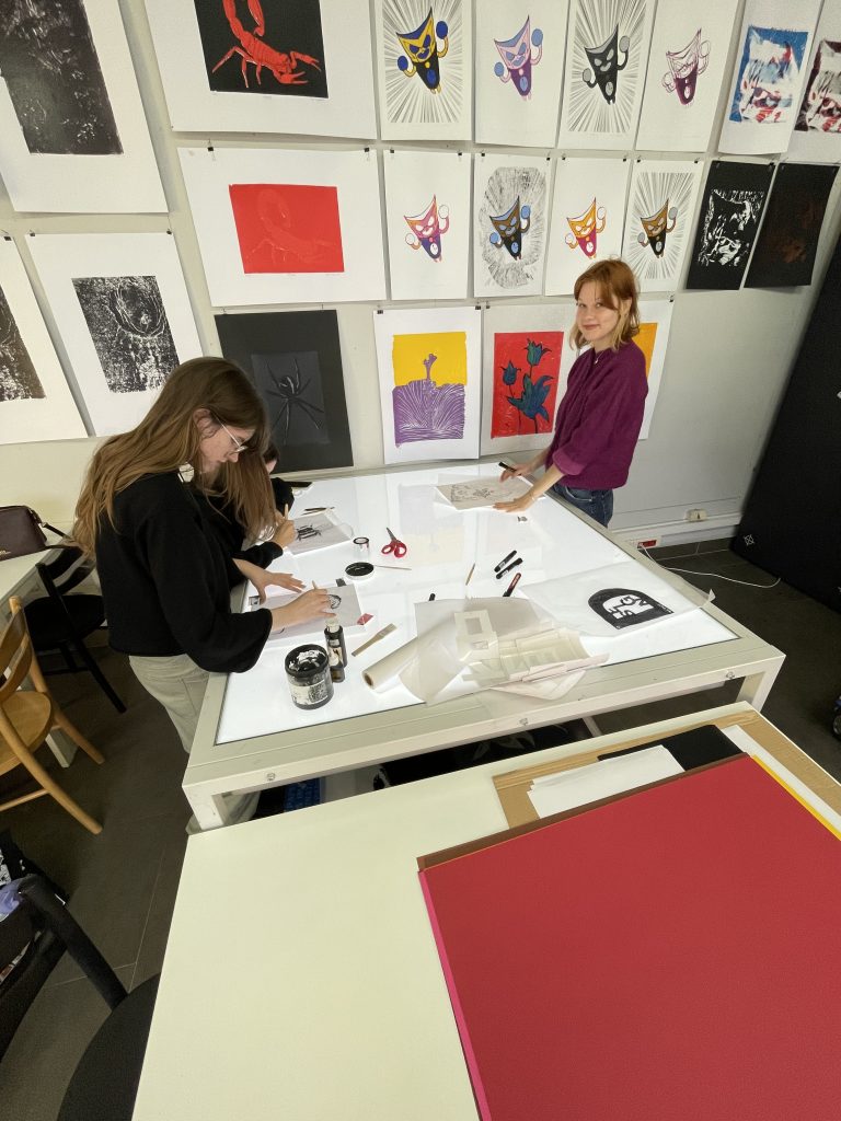 Widok z g&oacute;ry na duży st&oacute;ł podświetlany (podświetlarkę), przy kt&oacute;rym trzy osoby przygotowują projekty graficzne na kalkach. Ściana za nimi jest gęsto obwieszona pracami wykonanymi techniką sitodruku i linorytu, przedstawiającymi m.in. skorpiony i maski.