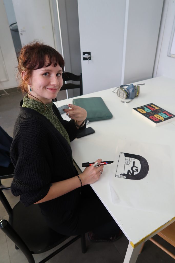 Młoda kobieta siedząca przy stole w pracowni, uśmiecha się do aparatu. W dłoni trzyma czarny marker, a przed nią leży szkic logotypu z napisem &bdquo;DESIGN&rdquo;. Na stole widoczne są r&oacute;wnież: smartfon, maska ochronna z filtrami oraz książka o typografii.