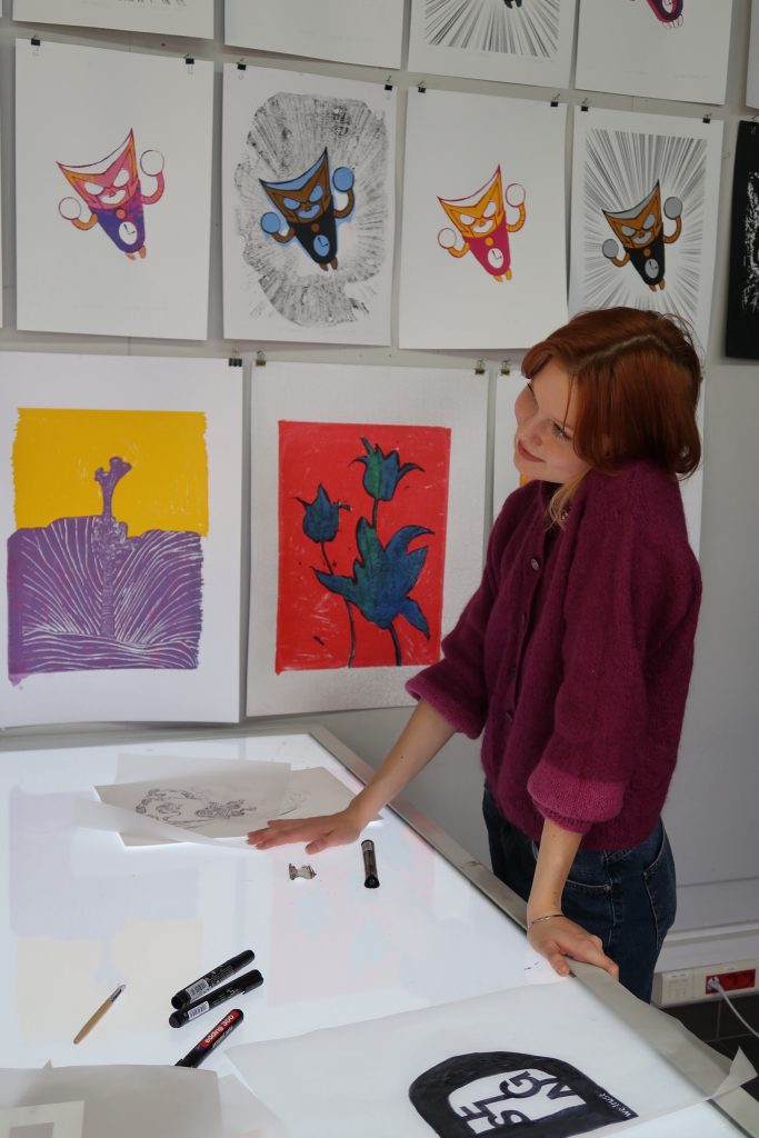 Zbliżenie na kobietę w bordowym swetrze stojącą przy stole podświetlanym. Kobieta patrzy na swoje projekty na kalkach. W tle widać kolorowe grafiki na ścianie, w tym motywy kwiatowe i ilustracje postaci.