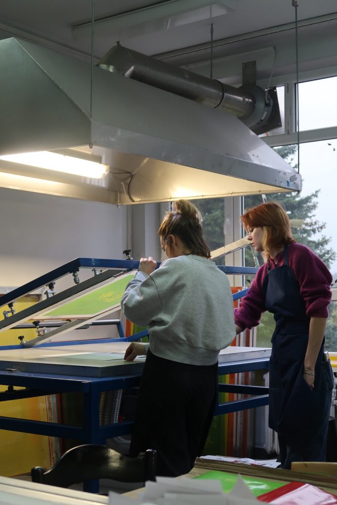 Dwie osoby pracują przy dużym stole do sitodruku pod metalowym okapem wentylacyjnym. Jedna z nich precyzyjnie ustawia papier pod uniesioną ramą sitodrukową.