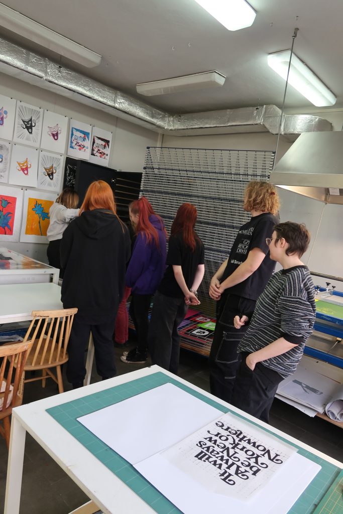 Grupa student&oacute;w w pracowni graficznej stoi przy regale do suszenia prac. Na pierwszym planie, na stole z zieloną matą do cięcia, leży wydruk z typograficznym napisem &bdquo;It will Always be New York or Nowhere&rdquo;. W tle ściana z licznymi kolorowymi grafikami.