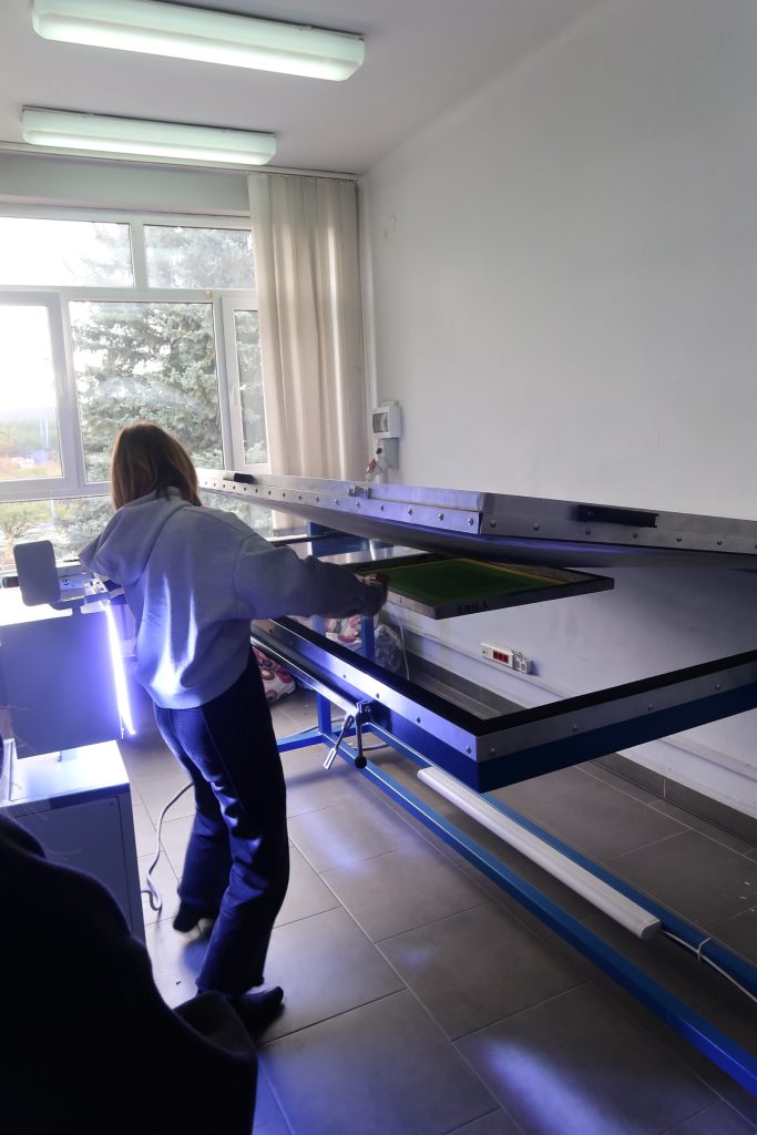 Osoba w bluzie z kapturem obsługuje duże urządzenie do naświetlania sit (kopioramę) w jasnym pomieszczeniu przy oknie. Urządzenie emituje niebieskie światło UV.