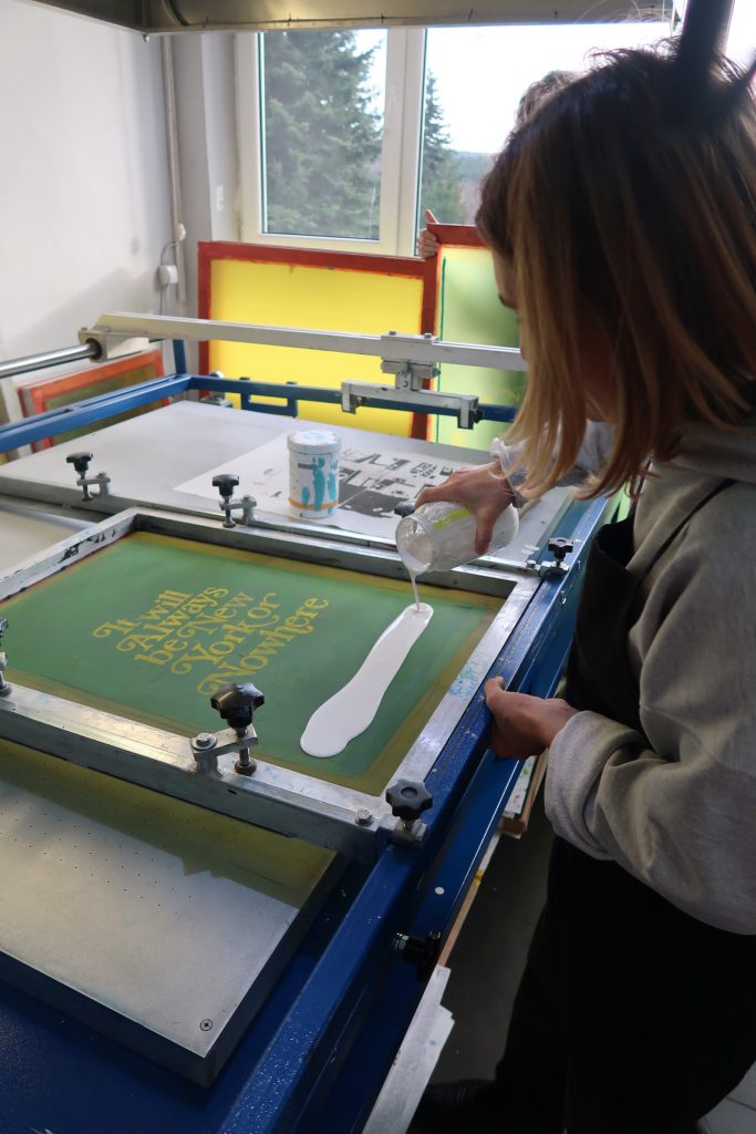 Zbliżenie na proces nakładania farby. Kobieta wylewa białą farbę z słoika na zieloną siatkę sitodrukową, na kt&oacute;rej widoczny jest szablon z napisem &bdquo;It will Always be New York or Nowhere&rdquo;