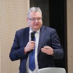 Mężczyzna w okularach, ciemnym garniturze i niebieskim krawacie przemawia do mikrofonu, trzymając w drugiej ręce pilot do prezentacji. Tło stanowią jasne, perforowane panele ścienne.