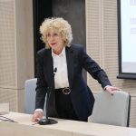 Rektor UJK prof. Beata Wojciechowska stoi przy stole prezydialnym, opierając się o krzesło. Ma krótkie, jasne, kręcone włosy i okulary. W tle widoczny fragment ekranu z tekstem.