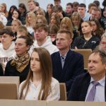 Uczestnicy wykładu siedzący w ławkach auli. Na pierwszym planie widać kobietę w białej koszuli oraz mężczyznę w garniturze, za nimi rzędy skupionych studentów.