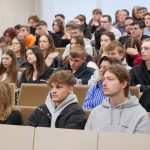 bliżenie na grupę studentek i studentów siedzących w auli. Młodzi ludzie w skupieniu słuchają wykładu; w kadrze widoczne są osoby w różnokolorowych bluzach i swetrach.