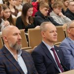 rzech mężczyzn w garniturach siedzi obok siebie w auli, obserwując przebieg wydarzenia. W tle, w nieostrości, widoczne są rzędy studentów.