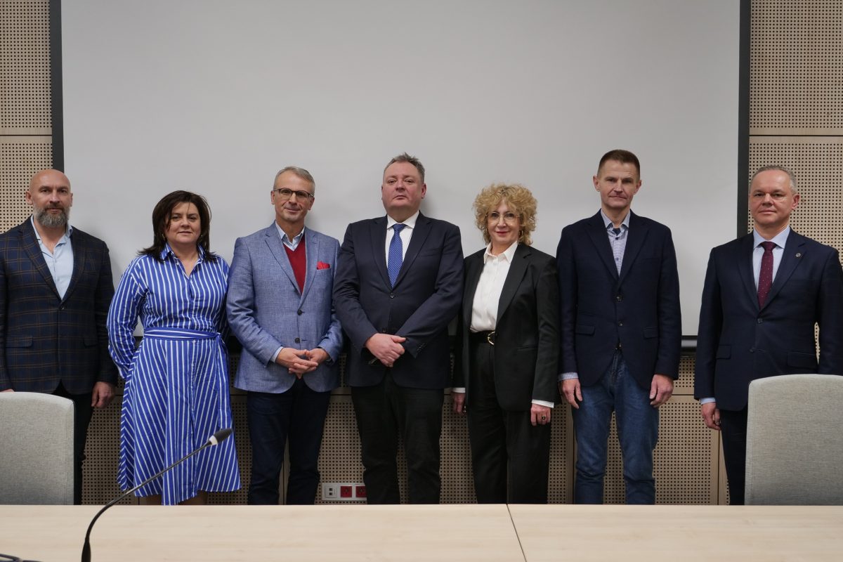 Pamiątkowe zdjęcie siedmiu osób stojących w rzędzie na tle białego ekranu. W centrum dr Piotr Szpunar oraz rektor prof. Beata Wojciechowska w otoczeniu pozostałych uczestników spotkania.