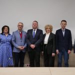 Pamiątkowe zdjęcie siedmiu osób stojących w rzędzie na tle białego ekranu. W centrum dr Piotr Szpunar oraz rektor prof. Beata Wojciechowska w otoczeniu pozostałych uczestników spotkania.