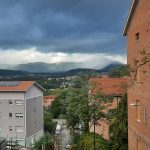 Widok z wzniesienia na miasto i g&oacute;ry pod ciemnymi, deszczowymi chmurami. Na pierwszym planie budynki z czerwonej cegły i dachy z dach&oacute;wki.