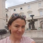 Selfie uśmiechniętej kobiety w r&oacute;żowej bluzce na tle zabytkowego placu z klasycystyczną fasadą budynku i kamienną fontanną.