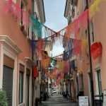 Wąska włoska uliczka ozdobiona kolorowymi girlandami z tiulu rozpiętymi między kamienicami. W oddali widoczne g&oacute;ry pod pochmurnym niebem.