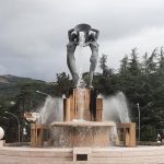 Monumentalna fontanna z rzeźbą dw&oacute;ch kobiet wylewających wodę z dużego naczynia. W tle gęste, ciemnozielone drzewa iglaste.