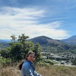 Kobieta na wzg&oacute;rzu z panoramą doliny, miasteczka i wysokich pasm g&oacute;rskich w tle pod błękitnym niebem z lekkimi chmurami.