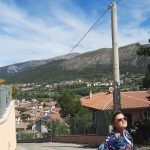 Kobieta w kolorowej kurtce stoi na stromej drodze prowadzącej w d&oacute;ł do miasteczka. W tle rozległy widok na g&oacute;ry i błękitne niebo.