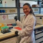 Kobieta w białym fartuchu i czarnych rękawiczkach pracuje w laboratorium, trzymając pipetę automatyczną przy statywie z prob&oacute;wkami.