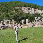 Kobieta w okularach przeciwsłonecznych stoi na tle antycznych, kamienno-ceglanych ruin amfiteatru u podn&oacute;ża zielonego, zalesionego wzg&oacute;rza.