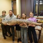 rupa os&oacute;b prezentuje duże, realistyczne pluszaki w kształcie ryb. Największy model, sum, jest trzymany przez kilka os&oacute;b na środku. Wszyscy uśmiechają się do obiektywu w jasnej sali wykładowej.