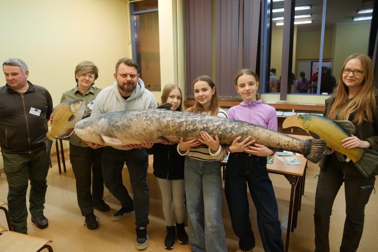 rupa osób prezentuje duże, realistyczne pluszaki w kształcie ryb. Największy model, sum, jest trzymany przez kilka osób na środku. Wszyscy uśmiechają się do obiektywu w jasnej sali wykładowej.