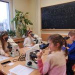 Dzieci w sali lekcyjnej patrzą przez mikroskopy ustawione na długim stole. Pomaga im starsza kobieta w okularach. W tle widać dużą roślinę doniczkową oraz czarną tablicę z kredowym napisem &bdquo;Noc Biolog&oacute;w 2026. Podr&oacute;ż w mikroskopijny świat&rdquo;.