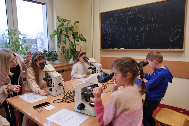 Dzieci w sali lekcyjnej patrzą przez mikroskopy ustawione na długim stole. Pomaga im starsza kobieta w okularach. W tle widać dużą roślinę doniczkową oraz czarną tablicę z kredowym napisem „Noc Biologów 2026. Podróż w mikroskopijny świat”.