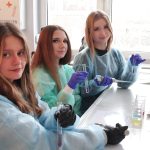 Trzy dziewczyny w kolorowych fartuchach ochronnych siedzą przy laboratoryjnym blacie. Każda trzyma prob&oacute;wkę z kolorowym płynem. Na stole widać pipetę automatyczną, zlewki z kolorowymi substancjami i stojaki na prob&oacute;wki.