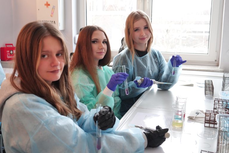 Trzy dziewczyny w kolorowych fartuchach ochronnych siedzą przy laboratoryjnym blacie. Każda trzyma probówkę z kolorowym płynem. Na stole widać pipetę automatyczną, zlewki z kolorowymi substancjami i stojaki na probówki.