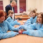 Cztery dziewczyny w błękitnych fartuchach ochronnych i rękawiczkach siedzą przy stole, wykonując sekcję serca wieprzowego. Używają metalowych narzędzi chirurgicznych. W tle stoi mężczyzna, a na szafce widać model anatomiczny ludzkiego oka.