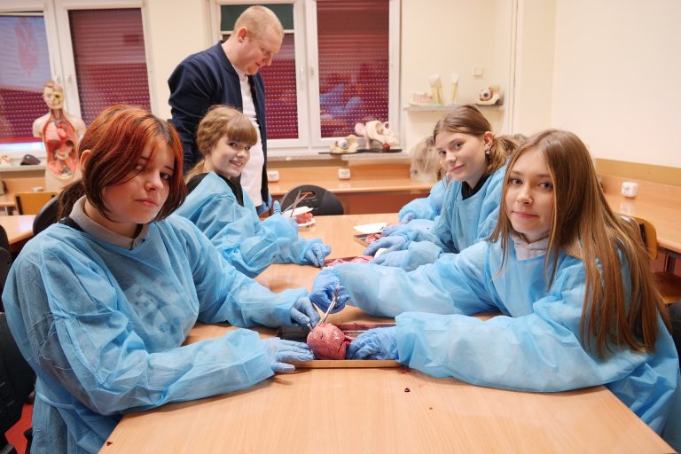 Cztery dziewczyny w błękitnych fartuchach ochronnych i rękawiczkach siedzą przy stole, wykonując sekcję serca wieprzowego. Używają metalowych narzędzi chirurgicznych. W tle stoi mężczyzna, a na szafce widać model anatomiczny ludzkiego oka.