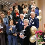 Uroczyste zgromadzenie gości i władz uczelni na schodach. Kobieta w niebieskim garniturze przemawia do mikrofonu. Wok&oacute;ł stoją osoby w eleganckich strojach z identyfikatorami. W tle widać choinkę i złote balony.