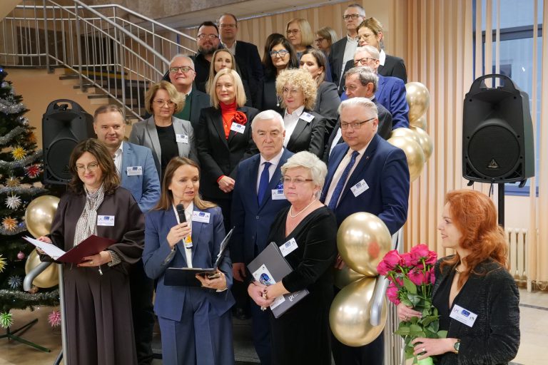 Uroczyste zgromadzenie gości i władz uczelni na schodach. Kobieta w niebieskim garniturze przemawia do mikrofonu. Wokół stoją osoby w eleganckich strojach z identyfikatorami. W tle widać choinkę i złote balony.