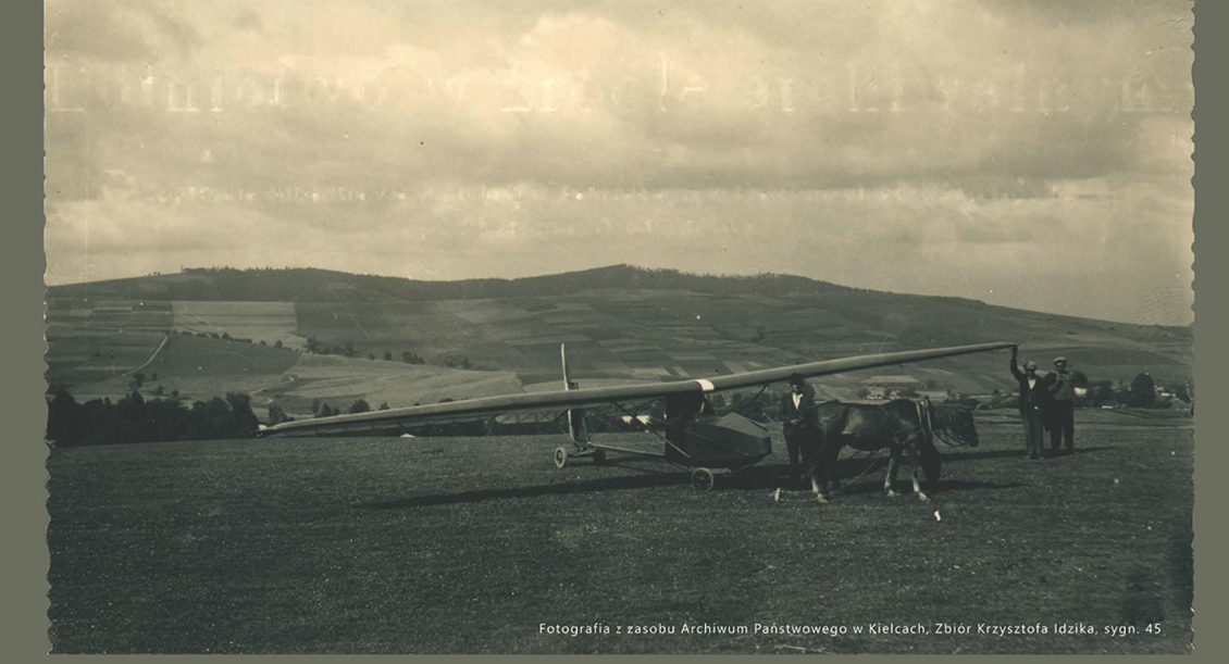 Archiwalna, czarno-biała fotografia przedstawiająca szybowiec na polanie na tle zalesionych wzgórz. Obok maszyny stoi koń oraz trzej mężczyźni; jeden z nich podtrzymuje skrzydło. Zdjęcie ma ząbkowane brzegi. U dołu widnieje napis informujący, że fotografia pochodzi z zasobów Archiwum Państwowego w Kielcach, ze zbioru Krzysztofa Idzika.