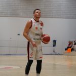 Mężczyzna w białym stroju sportowym z numerem 13 stoi na parkiecie, trzymając piłkę do koszyk&oacute;wki. Patrzy w g&oacute;rę, skupiony przed rzutem. W tle widać siatkę ochronną i widownię na pomarańczowych krzesłach.