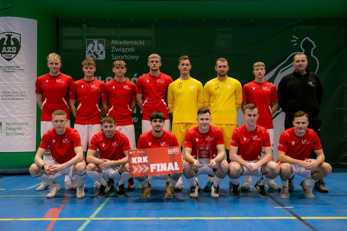 Grupowe zdjęcie męskiej drużyny futsalu na niebieskiej hali. Zawodnicy w czerwonych i żółtych koszulkach z logo „UJK”. Dwaj klęczący z przodu trzymają tablicę z napisem „KRK FINAL” oraz statuetkę. W tle banery „AZS”.