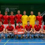 Grupowe zdjęcie męskiej drużyny futsalu na niebieskiej hali. Zawodnicy w czerwonych i ż&oacute;łtych koszulkach z logo &bdquo;UJK&rdquo;. Dwaj klęczący z przodu trzymają tablicę z napisem &bdquo;KRK FINAL&rdquo; oraz statuetkę. W tle banery &bdquo;AZS&rdquo;.