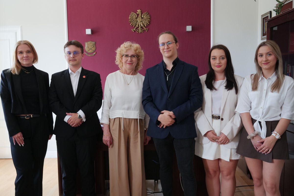 Szóstka uśmiechniętych osób – cztery kobiety i dwóch mężczyzn – stoi obok siebie w eleganckich strojach. Pozują w biurze na tle bordowej ściany, na której wisi godło Polski.