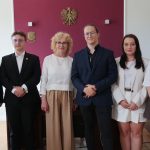 Szóstka uśmiechniętych osób – cztery kobiety i dwóch mężczyzn – stoi obok siebie w eleganckich strojach. Pozują w biurze na tle bordowej ściany, na której wisi godło Polski.