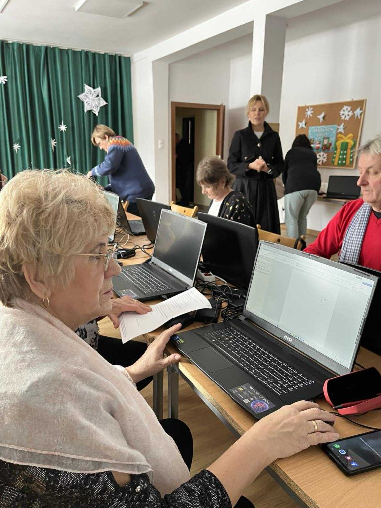 Uczestnicy (seniorzy) z gminy Masł&oacute;w podczas zajęć cyfrowych pracują przy laptopach pod nadzorem trenera UJK - 2