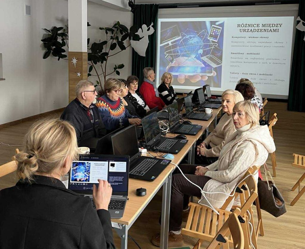 Uczestnicy (seniorzy) z gminy Masł&oacute;w podczas zajęć cyfrowych pracują przy laptopach pod nadzorem trenera UJK - 5