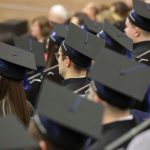 Grupa absolwentów w czarnych biretach i togach siedząca rzędami na auli podczas uroczystości.