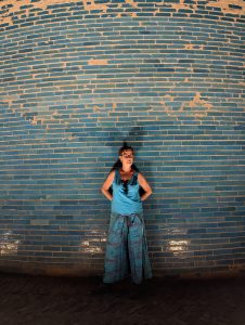 Osoba w turkusowej bluzce i sp&oacute;dnicy stoi pod p&oacute;łkolistą, niebieską ceglaną ścianą, ręce za plecami, patrzy w bok.