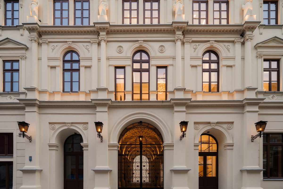 Fasada zabytkowej kamienicy Teatru Żeromskiego w jasnym piaskowcu. Na parterze ozdobna, kuta brama i stylowe latarnie. Wyżej rzędy łukowych okien z ciepłym światłem, rozdzielone korynckimi kolumnami. Na najwyższym piętrze, między oknami, cztery białe rzeźby przedstawiające postacie kobiece.