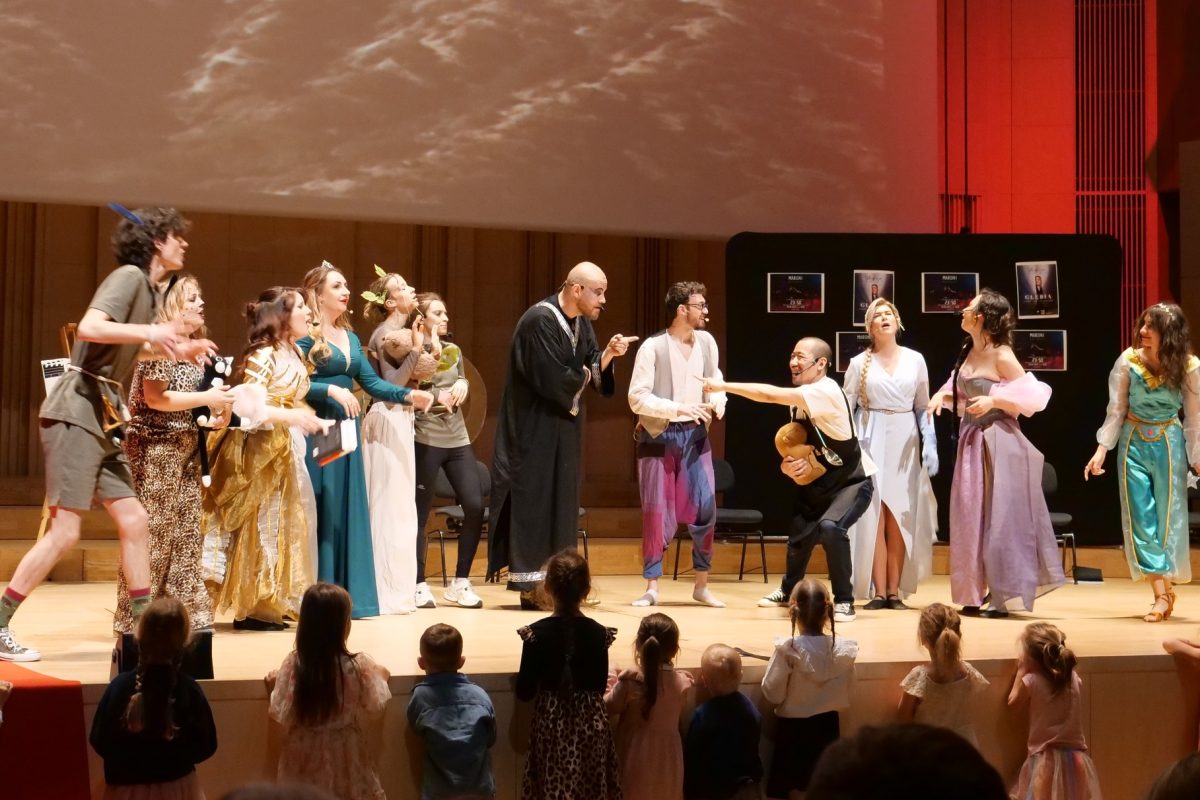 Scena ze spektaklu teatralnego dla dzieci. Grupa aktorów w różnorodnych, kolorowych kostiumach (m.in. suknie balowe, strój pantery, szaty) dynamicznie gestykuluje i śpiewa. W centrum łysy mężczyzna w czarnej todze. Przed podwyższoną sceną stoi rząd dzieci tyłem do obiektywu, z fascynacją oglądających przedstawienie. W tle widoczne plakaty.