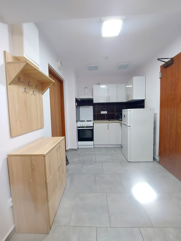 Zdjęcie przedstawia jasną kuchnię w Domu Studenta Asystent. W pomieszczeniu znajduje się zlewozmywak, płyta grzewcza z piekarnikiem, mała lod&oacute;wka oraz białe szafki kuchenne. Po lewej stronie wisi wieszak na ubrania, pod nim stoi szafka na buty. Kuchnia jest czysta i dobrze oświetlona.