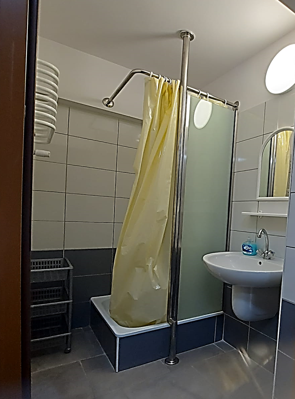 Zdjęcie przedstawia łazienkę Domu Studenta Asystent. Wnętrze wyłożone jest białymi oraz szarymi płytkami. W pomieszczeniu znajduje się prysznic z ż&oacute;łtą zasłoną prysznicową, umywalka z lustrem nad nią oraz szary plastikowy stojak z p&oacute;łkami. Łazienka jest czysta, dobrze oświetlona i urządzona w prostym, funkcjonalnym stylu.