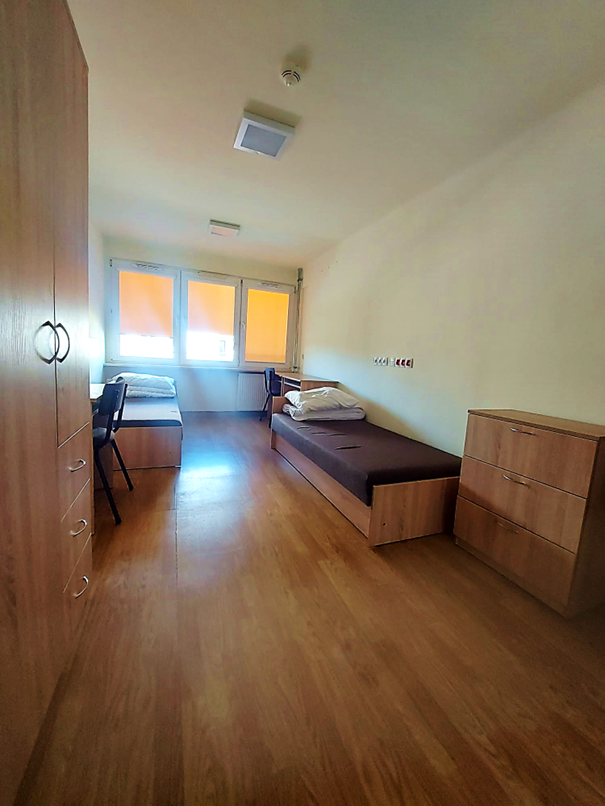 Zdjęcie przedstawia pok&oacute;j dwuosobowy w Domu Studenta Asystent. W pomieszczeniu znajdują się dwa ł&oacute;żka z białą kołdrą i poduszkami , dwa biurka, krzesła Pok&oacute;j jest jasny, z brązową podłogą i drewnianymi meblami.