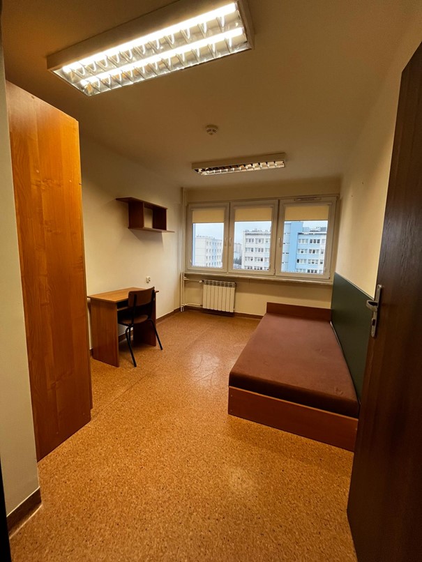 Jednoosobowy pok&oacute;j w domu studenta FAMA. W pomieszczeniu znajduje się proste wyposażenie: po prawej stronie stoi ł&oacute;żko z brązowym materacem, a po lewej biurko z czarnym krzesłem. Nad biurkiem umieszczona jest mała wisząca p&oacute;łka. Na końcu pokoju znajdują się trzy duże okna z widokiem na pobliskie bloki mieszkalne. Całość oświetlają dwie sufitowe lampy jarzeniowe.